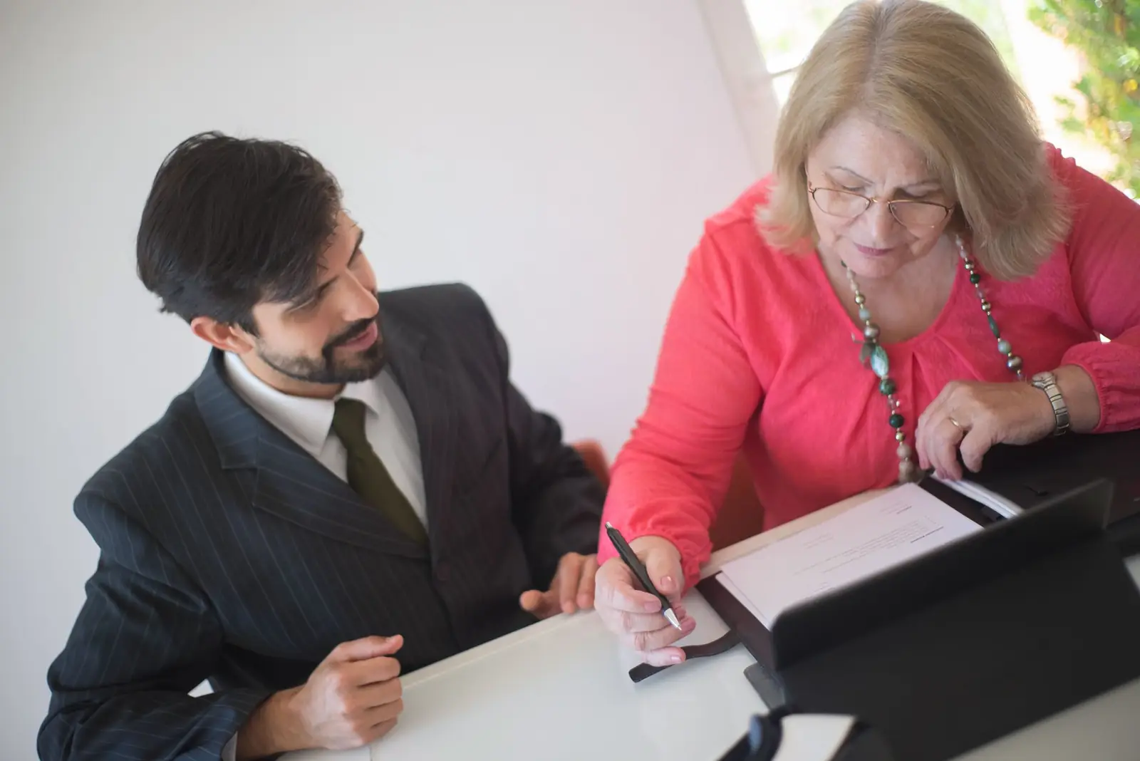 A man in a suit guiding an older woman as she signs a document at a table